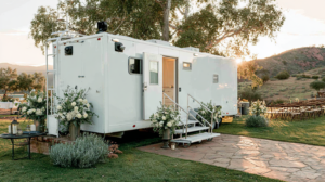 A large white mobile trailer with steps and an open door, decorated with white flower arrangements, is parked on grass with trees and outdoor seating nearby, lit by sunlight.