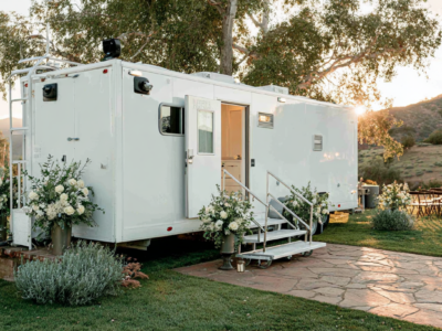 A large white mobile trailer with steps and an open door, decorated with white flower arrangements, is parked on grass with trees and outdoor seating nearby, lit by sunlight.