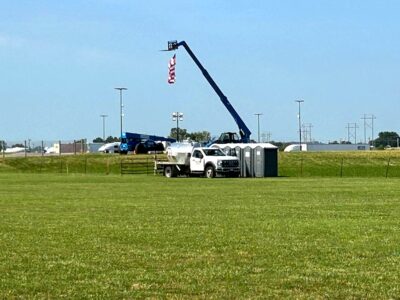 A white utility truck is parked on grass near portable toilets. Behind it, a blue crane lifts an American flag. Industrial buildings and light poles are visible in the background under a clear blue sky.