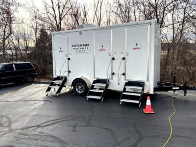 A mobile restroom trailer with three doors, marked for men and women, is parked on asphalt near trees. Steps lead to each door, with a traffic cone and a yellow extension cord nearby. A black SUV is parked nearby.