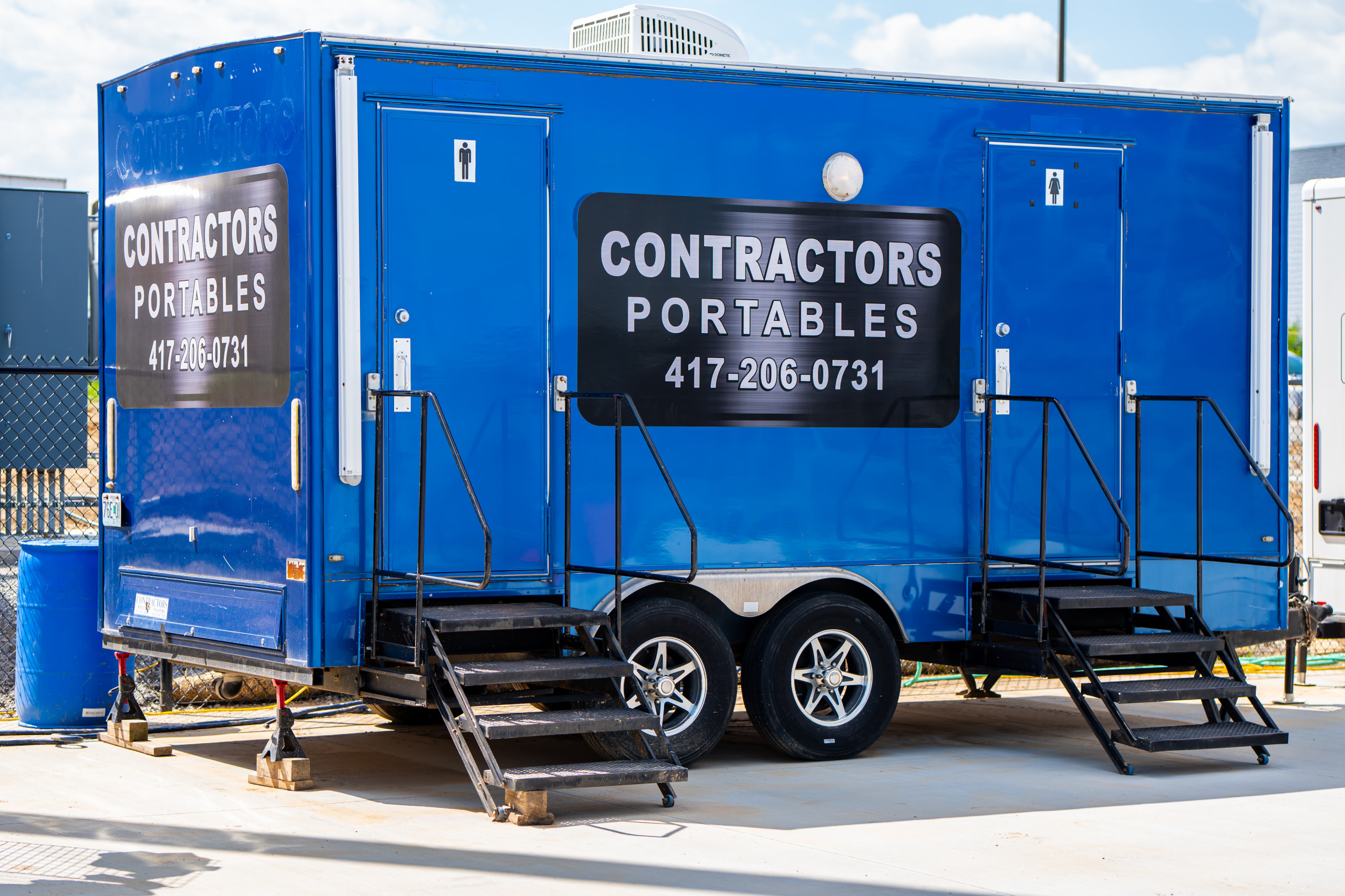Bright blue portable restroom trailer with two staircases and labeled “Contractors Portables” along with a phone number, parked on pavement near a fence on a sunny day.