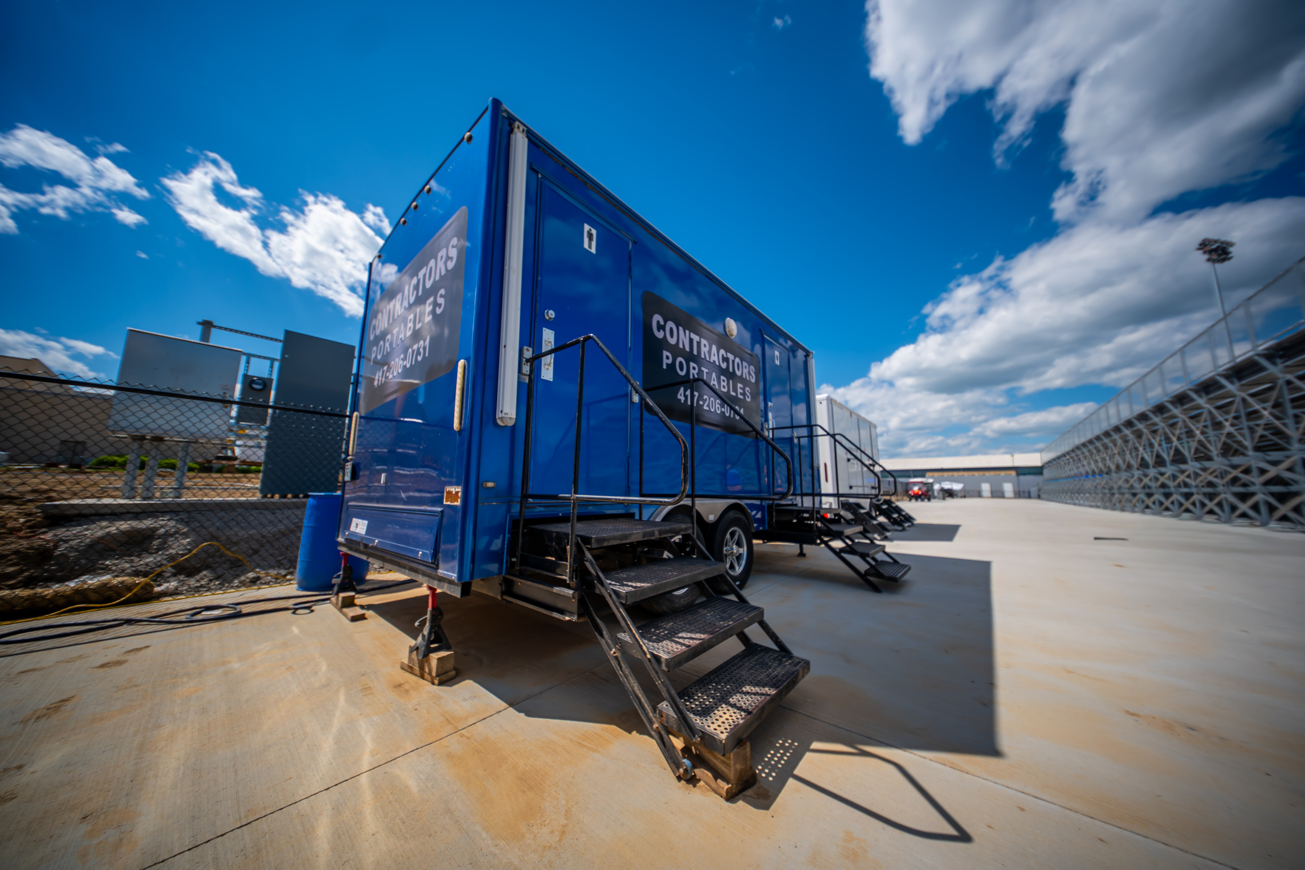 A blue portable restroom trailer with metal stairs is parked on a concrete surface near a chain-link fence, under a partly cloudy sky.