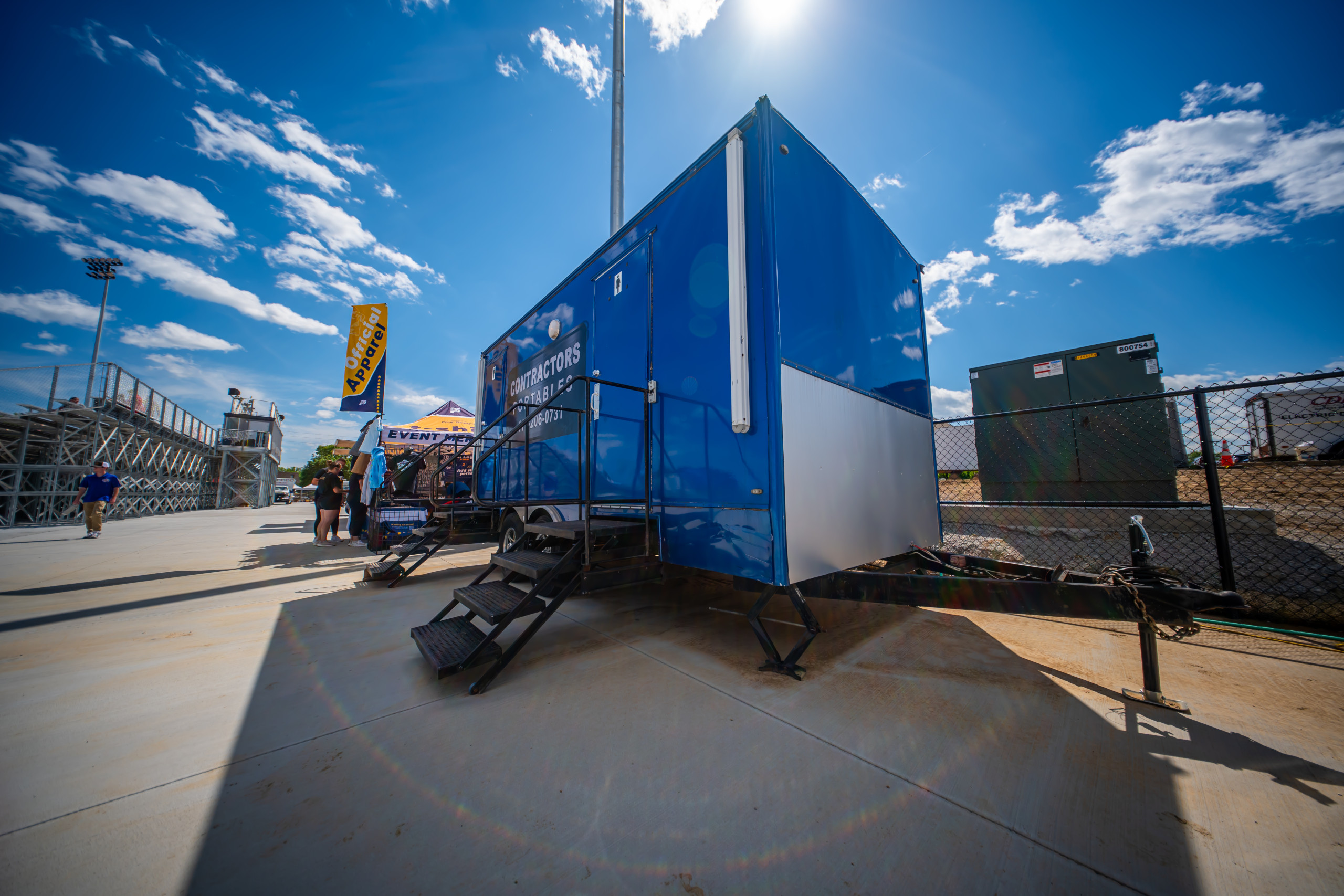 A bright blue mobile trailer with stairs is parked on a sunny day near a fenced area, with a blue sky and scattered clouds above. People and a tent are visible in the background near metal bleachers.