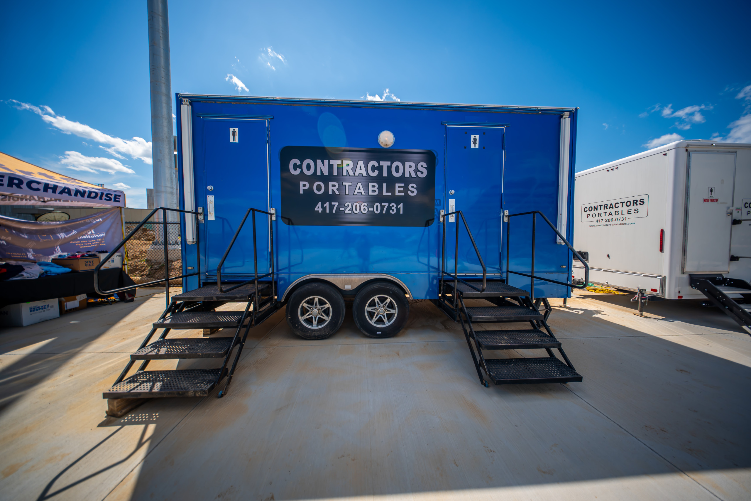 A blue portable restroom trailer with two sets of stairs, labeled "Contractors Portables" and a phone number, is parked on concrete under a clear blue sky.