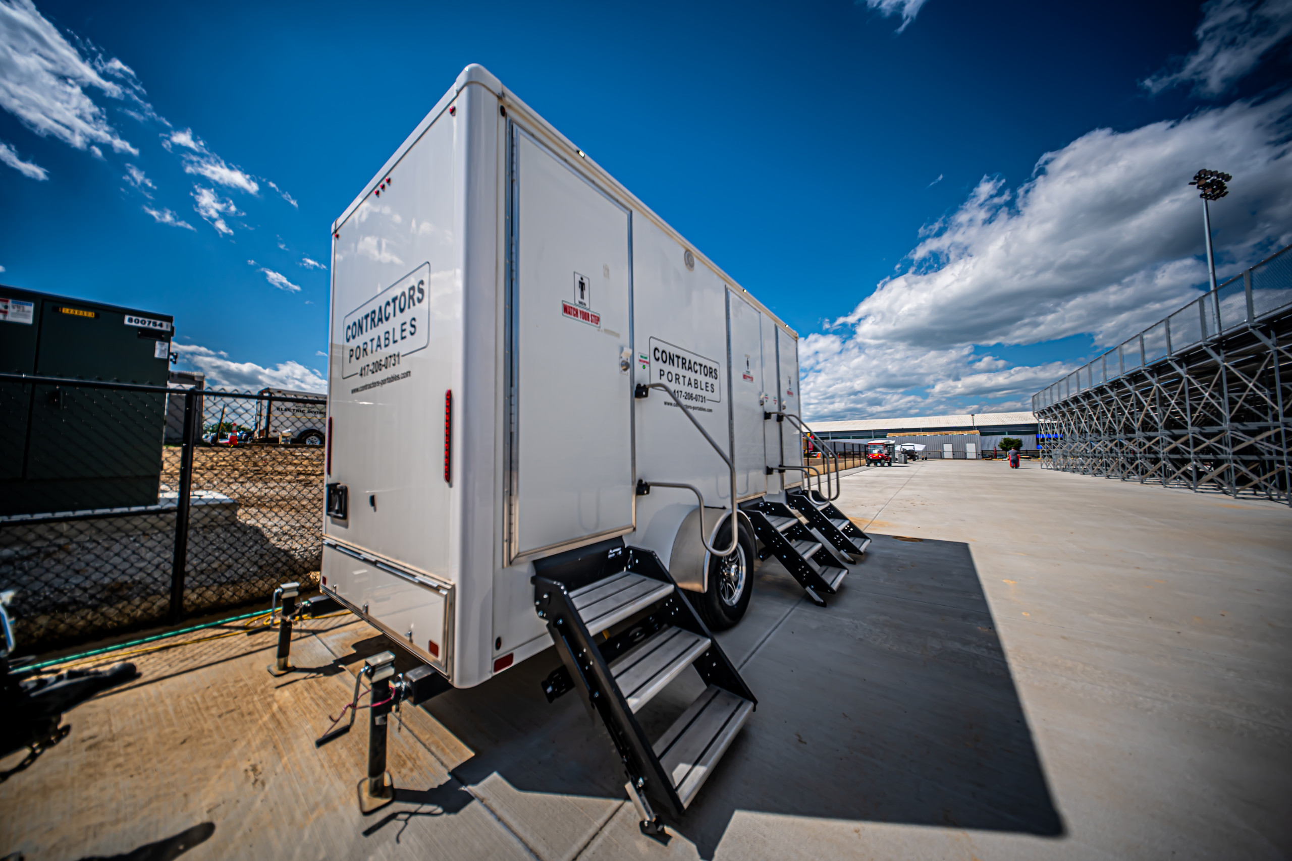 A white portable restroom trailer with steps is parked on a paved area near a chain-link fence, with a blue sky and clouds overhead. Signs on the trailer read "Contractors Portables.