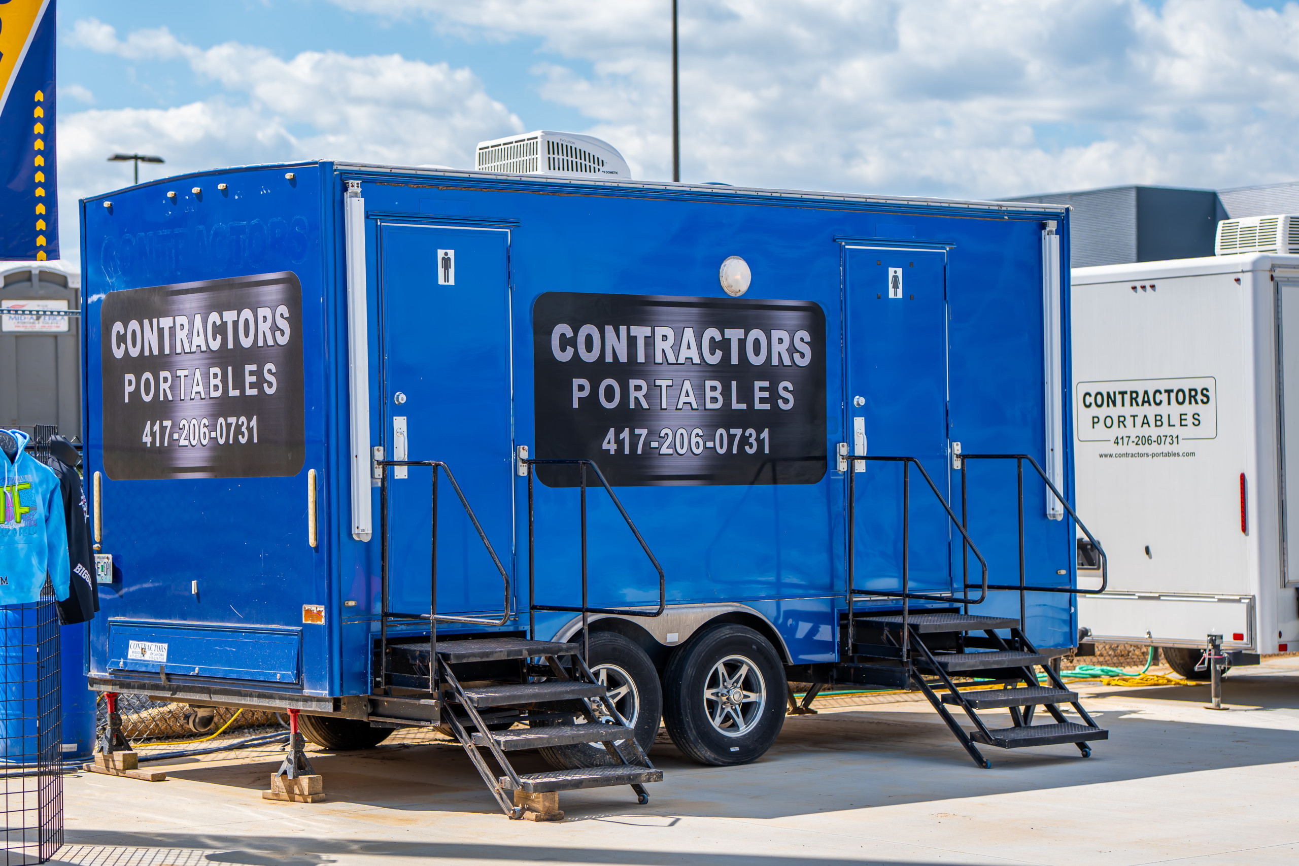 A bright blue portable restroom trailer with stairs and "Contractors Portables" signage is parked on a paved lot under a partly cloudy sky. Another white contractor portable unit is visible in the background.