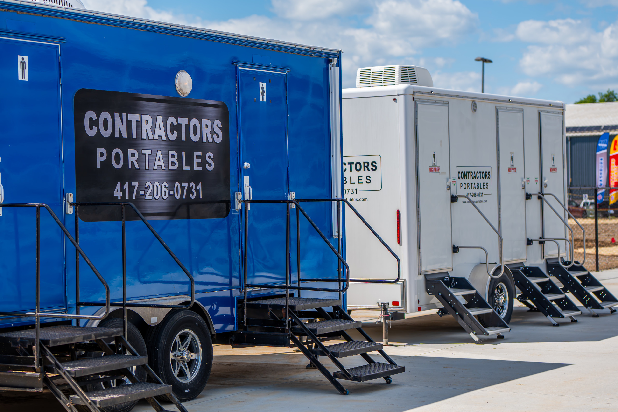 Several portable restroom trailers parked on a paved surface, with black steps leading up to each entrance. The blue trailer displays "CONTRACTORS PORTABLES" and a phone number. The sky is partly cloudy.