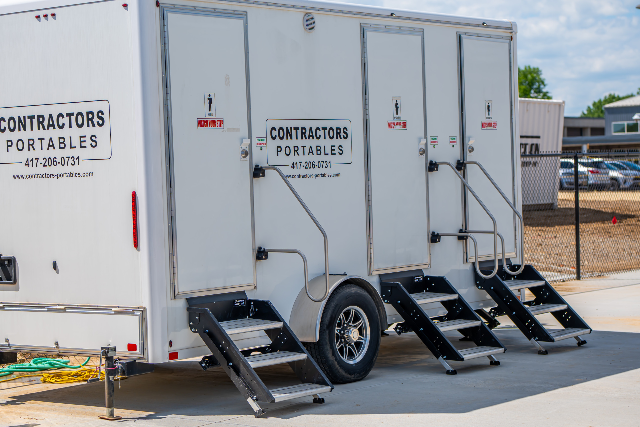 A row of three portable restroom trailers labeled "Contractors Portables" with black stairs and railings, parked on a paved surface near a fenced area and some buildings.