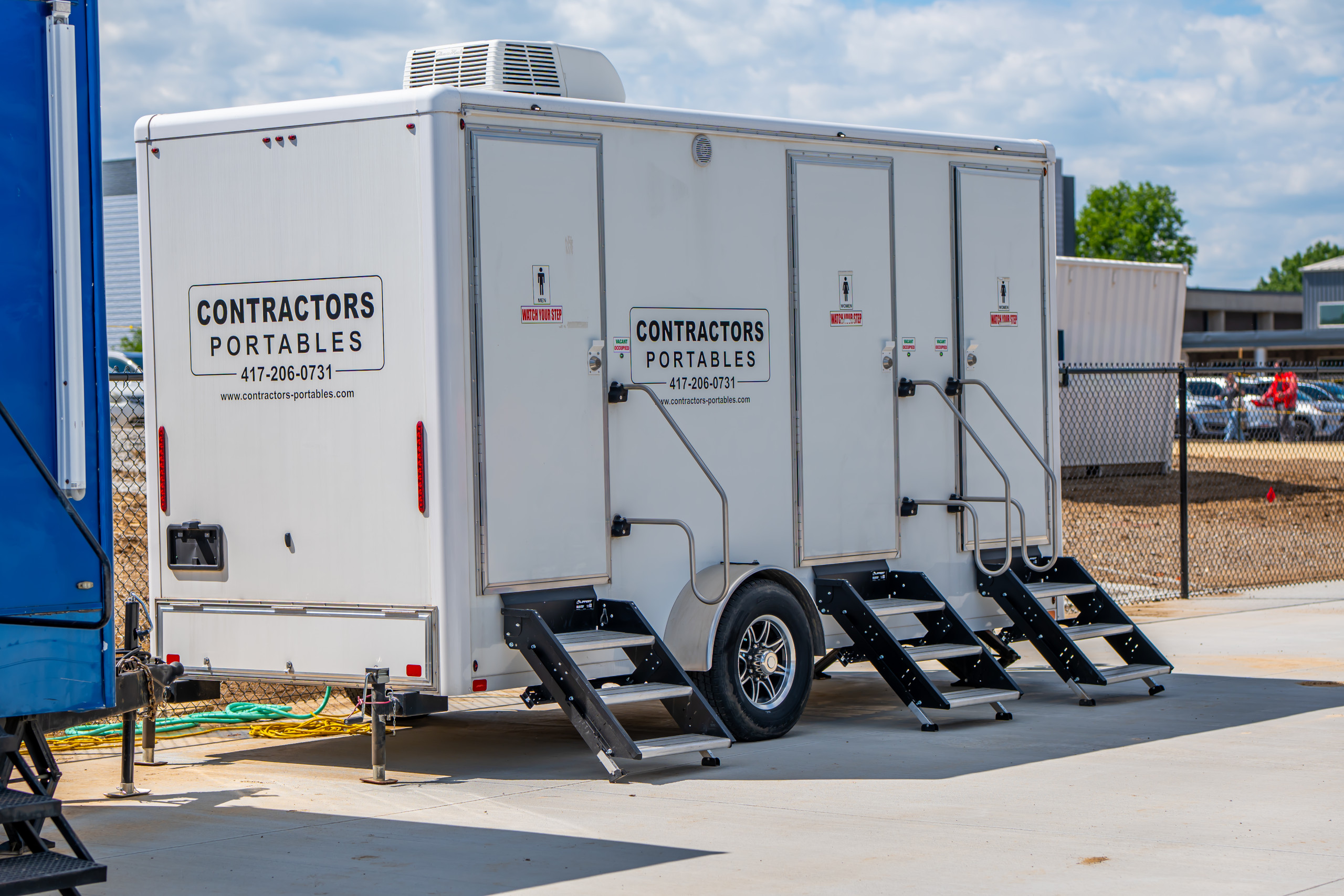 A white portable restroom trailer with three separate doors and steps, labeled "Contractors Portables," is parked on concrete near a construction site on a sunny day.
