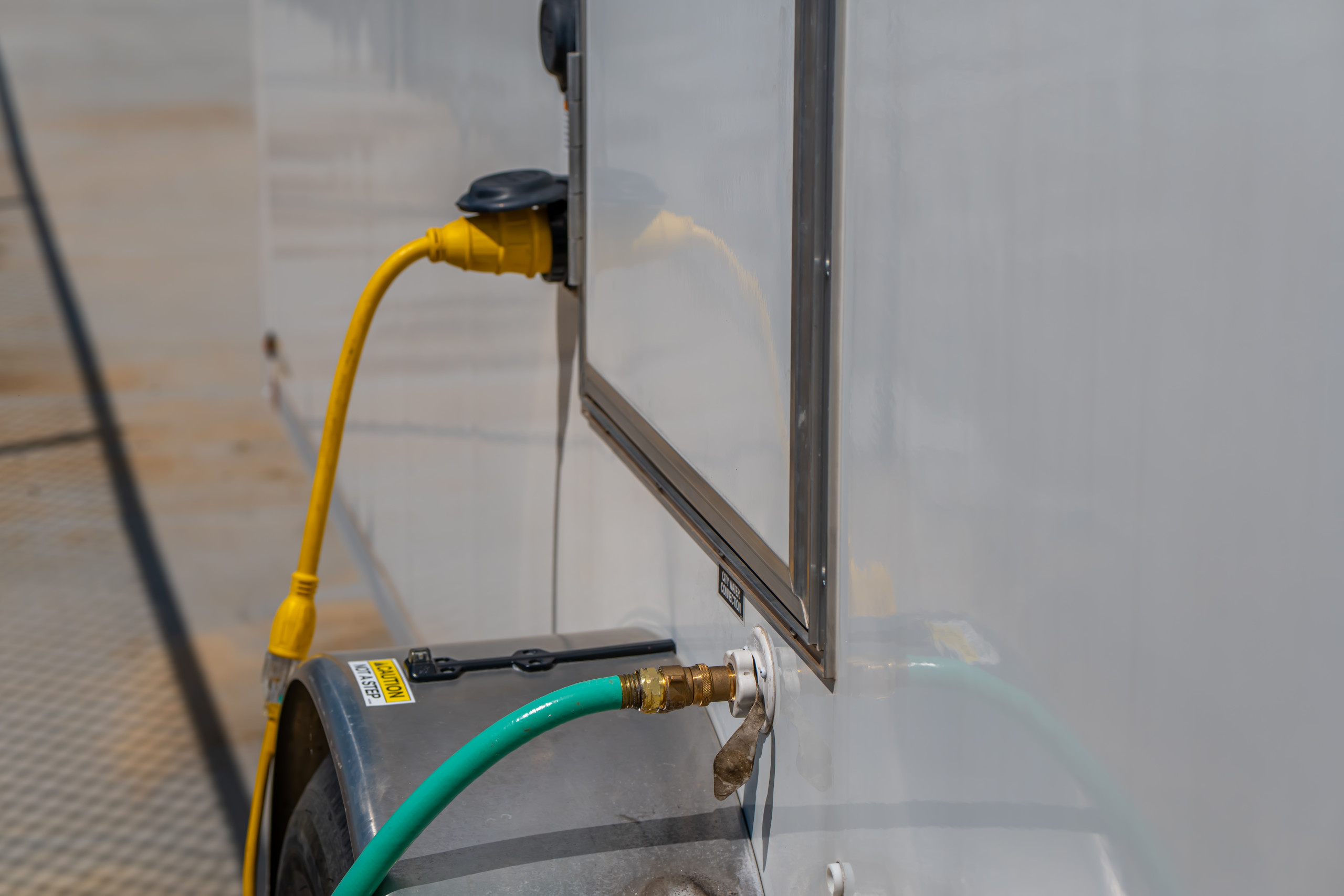 A close-up of an RV utility hookup shows a yellow electrical cable plugged into a socket and a green hose connected to a water inlet on the side of the vehicle.