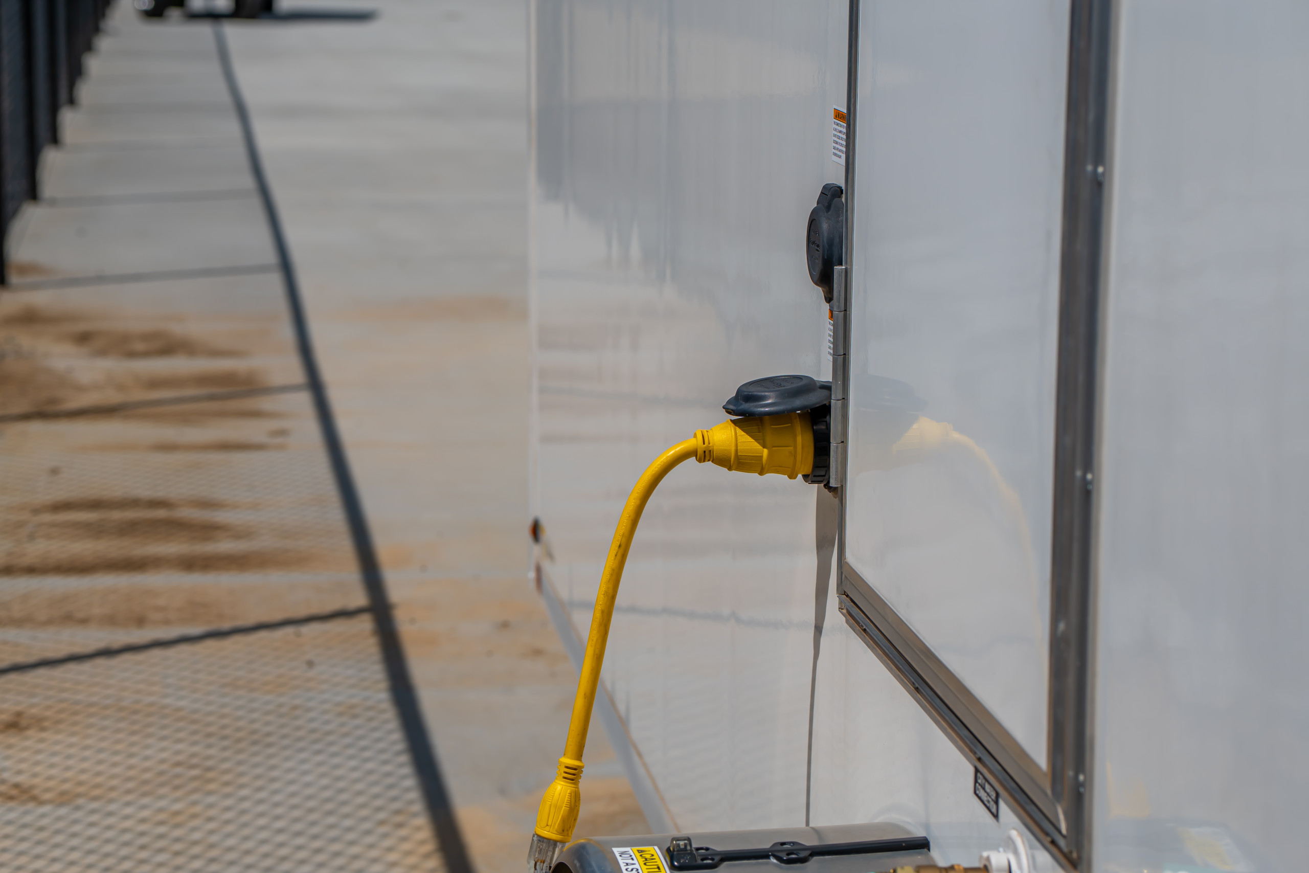 A bright yellow power cable is plugged into the side of a white trailer parked on a concrete surface. The surroundings show the trailer’s lower edge and a textured metal ramp.
