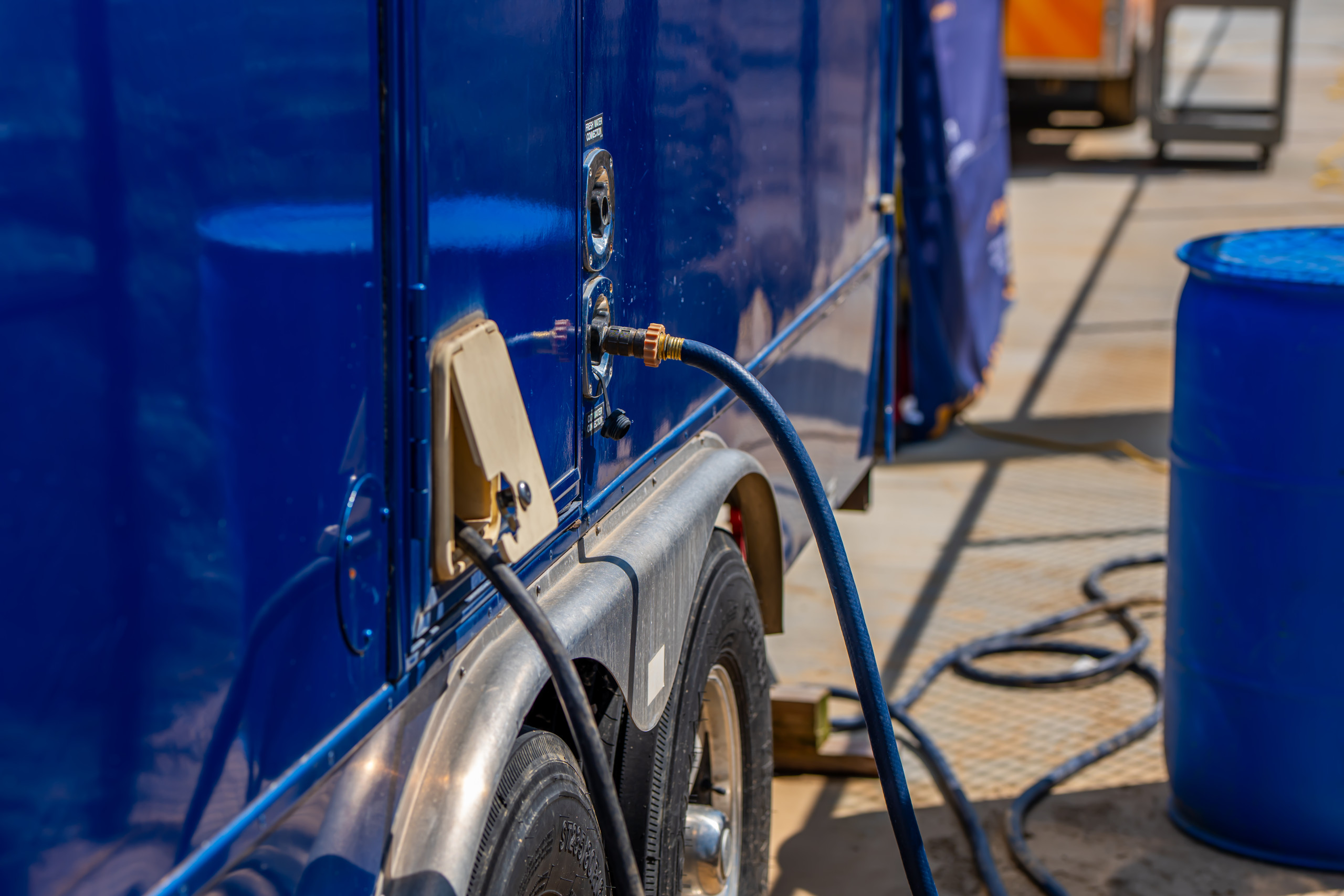 A close-up of a blue trailer with a hose connected to its side, parked on a paved surface. A blue barrel and a coiled hose are visible nearby in the background.