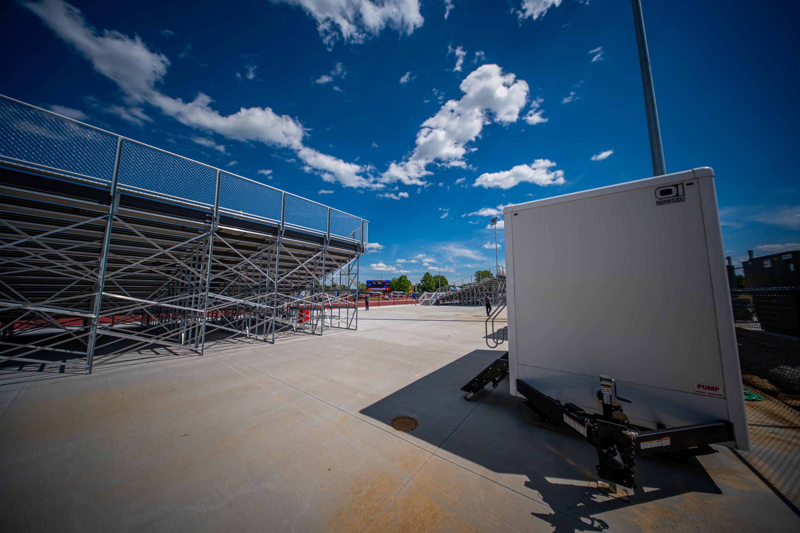A metal stadium bleacher is seen on the left under a blue sky with scattered clouds. To the right, a white portable trailer is parked on a wide, empty concrete area.