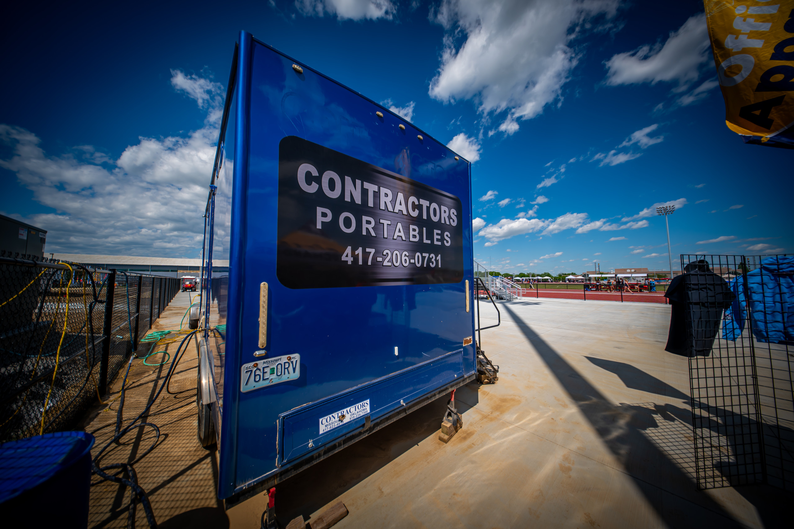 A blue portable restroom trailer labeled "CONTRACTORS PORTABLES 417-206-0731" sits on a paved area under a bright sky with clouds; a fence and sports field are visible in the background.
