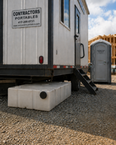 A portable contractor's office trailer with a sign, a white water tank underneath, and a portable toilet nearby, set on a gravel lot with wooden construction framing visible in the background.