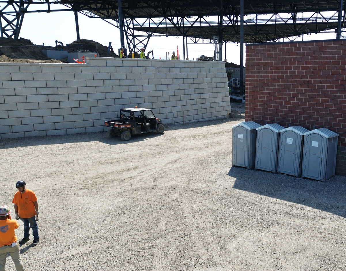 A construction site with two workers in orange vests, a utility vehicle on gravel, four portable toilets, a brick wall, and a partially built structure with scaffolding in the background.
