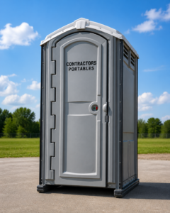 A single gray portable toilet labeled "Contractors Portables" stands on a paved area outdoors, with green grass, trees, and a blue sky with clouds in the background.