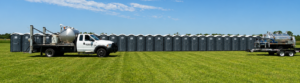 A row of portable toilets lines the edge of a grassy field under a blue sky. Two white trucks, one with a large tank, are parked in front of the toilets, likely servicing them.