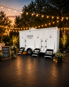 A white portable restroom trailer with three doors is set up outdoors at night, surrounded by string lights and trees. A wooden welcome sign and floral decorations are placed nearby on the pavement.